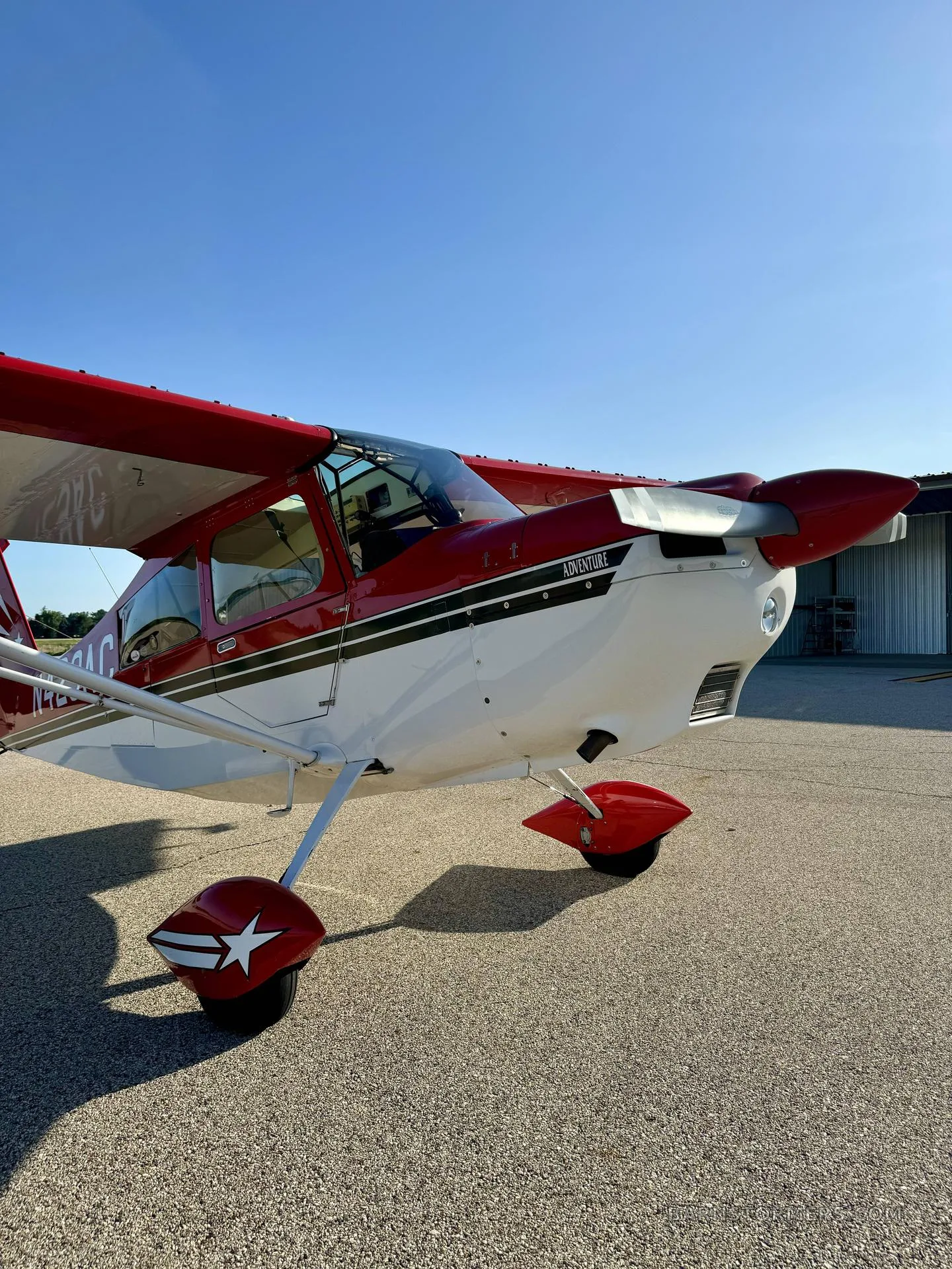 A close-up front view of the red and white Adventure aircraft, N429AC, with a star on the wheel fairings, parked on an airfield.