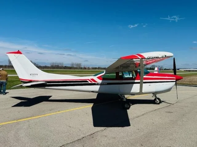 A white and red Cessna 172 aircraft with tail number N737FR is parked on an asphalt tarmac under a clear blue sky, with a person standing nearby.