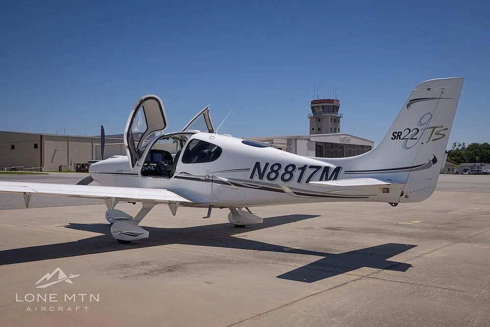 A rear-side view of a white Cirrus SR22T aircraft (N8817M) with doors open, parked on a tarmac.