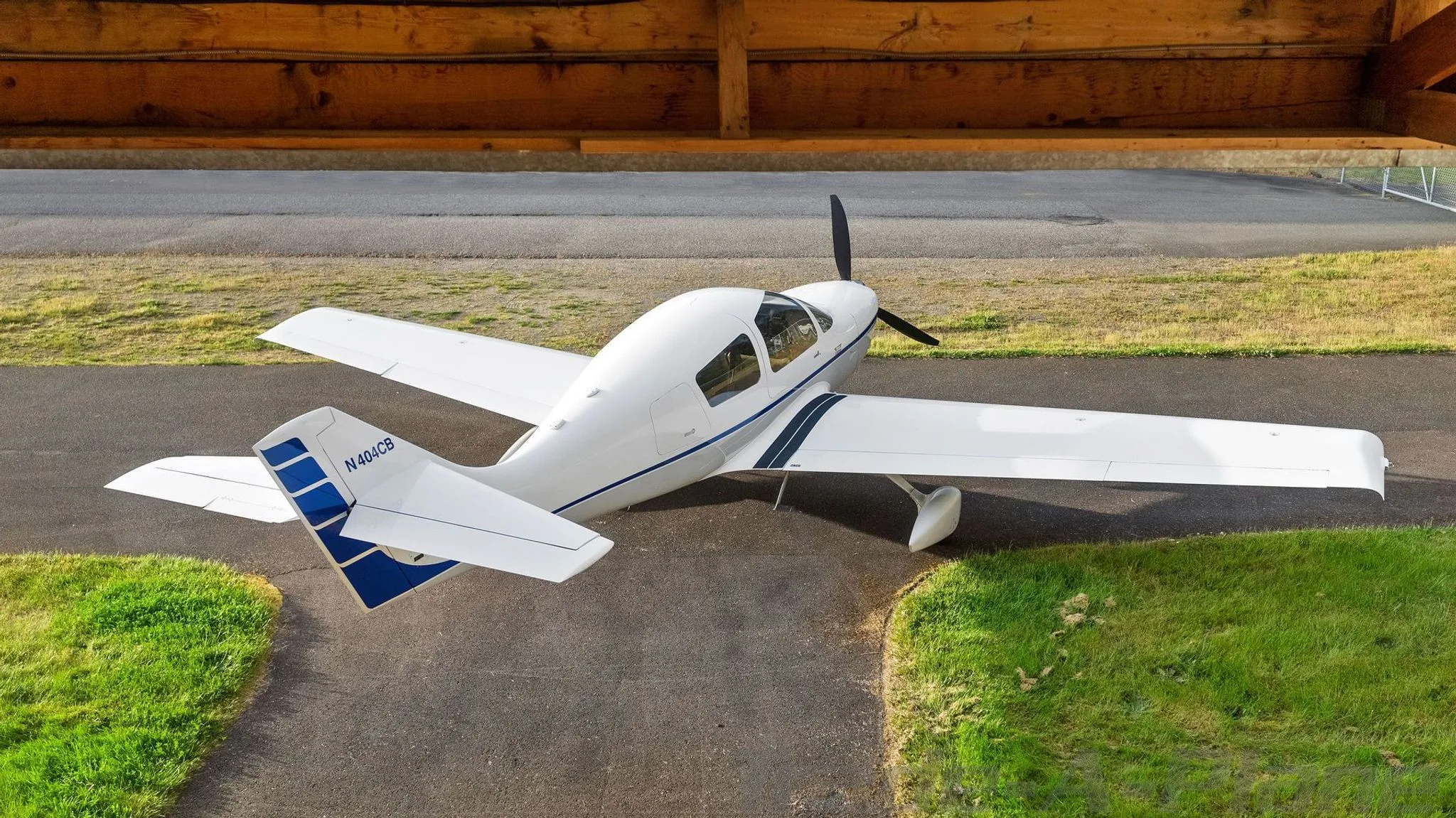 An overhead view of a white single-engine aircraft with blue stripes on the tail, registration N404CB, parked on an asphalt surface next to a wooden structure.