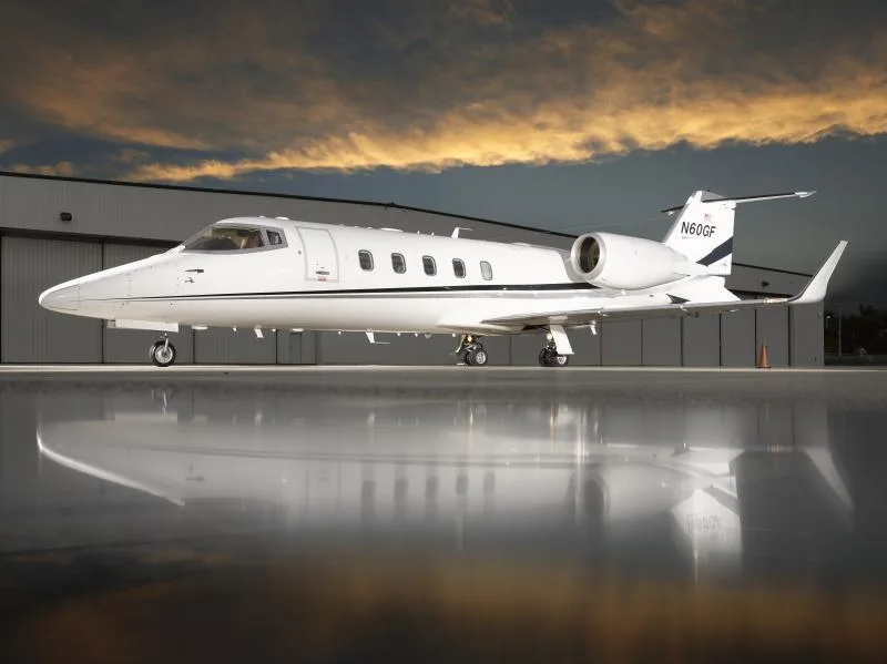 A white Learjet 60, registration N60GF, with black stripes, parked on a wet tarmac in front of a hangar at sunset.