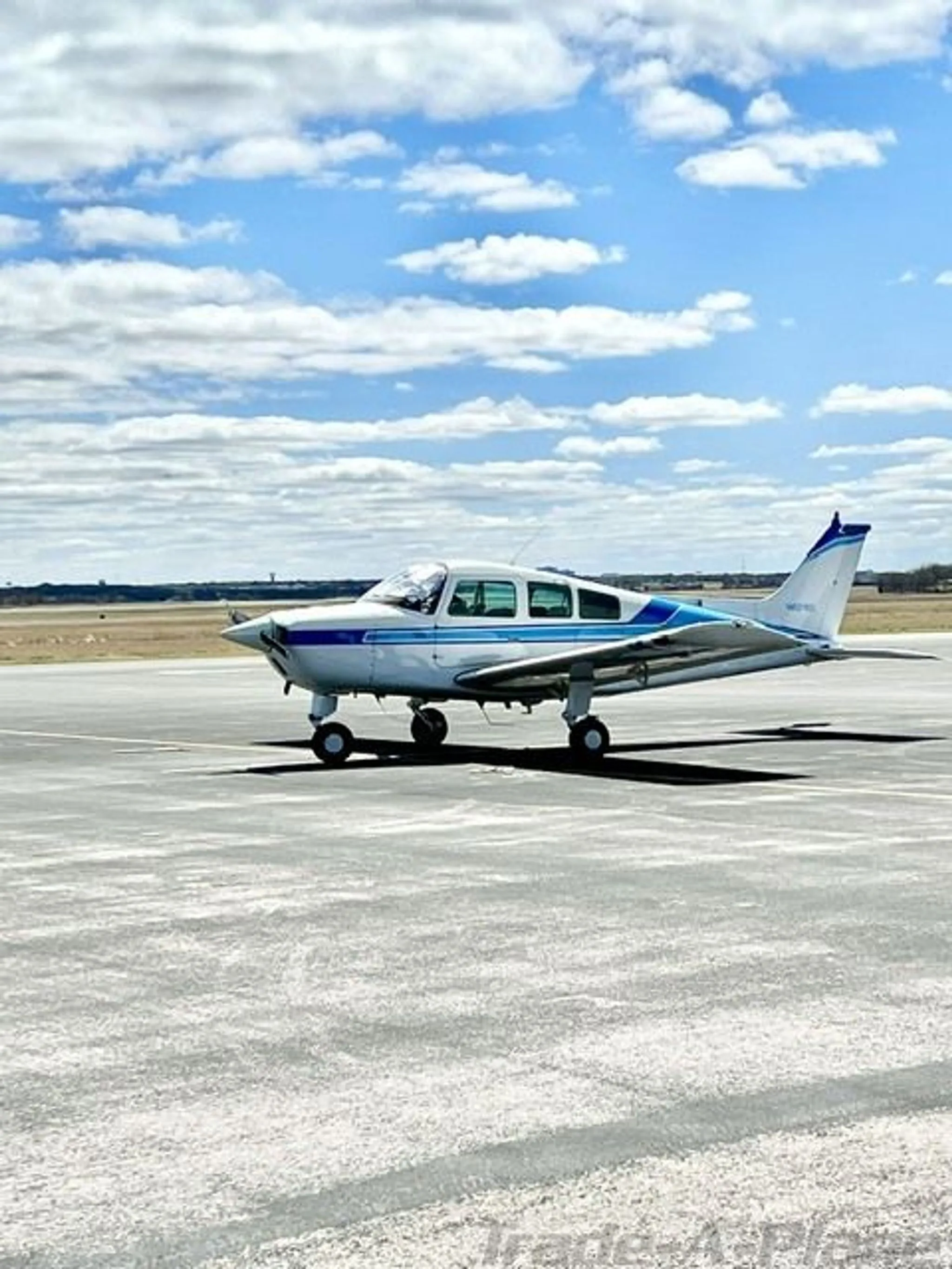 A white Beechcraft Sundowner N6718T with blue stripes parked on a tarmac under a partly cloudy sky, viewed from the side.
