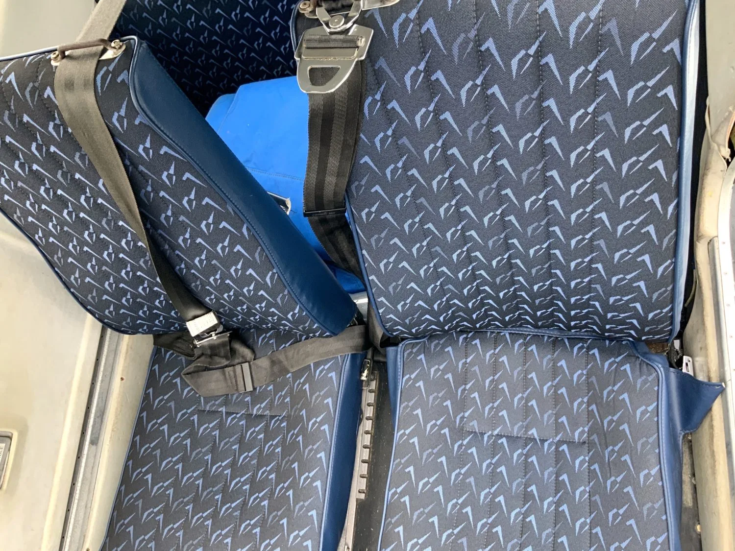 Two aircraft cabin seats with dark blue fabric and a light blue geometric pattern, with seatbelts fastened.