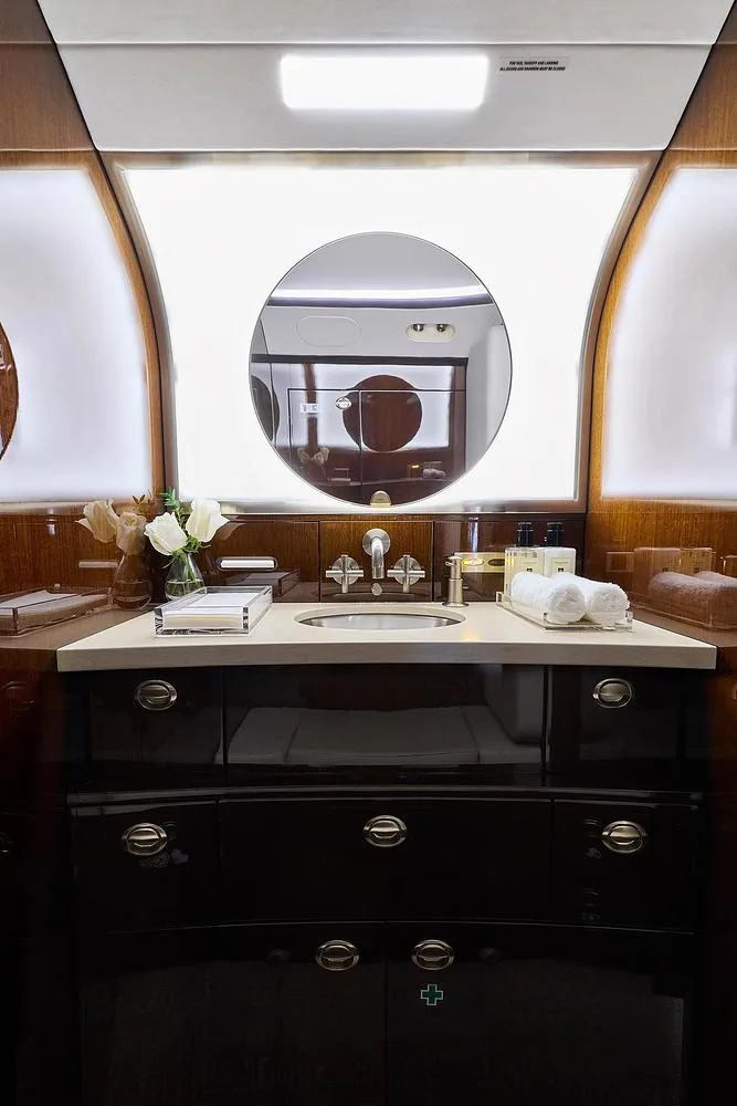 The elegant lavatory vanity of a business jet, featuring a sink, mirror, and dark wood cabinetry with white towels and flowers.