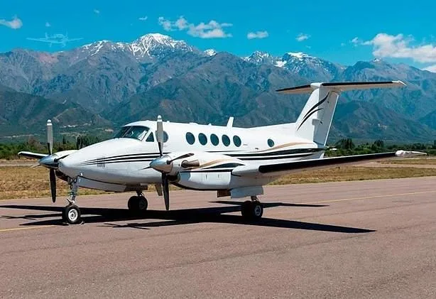 A white Beechcraft King Air turboprop aircraft with black and gold stripes is parked on a tarmac with mountains in the background.
