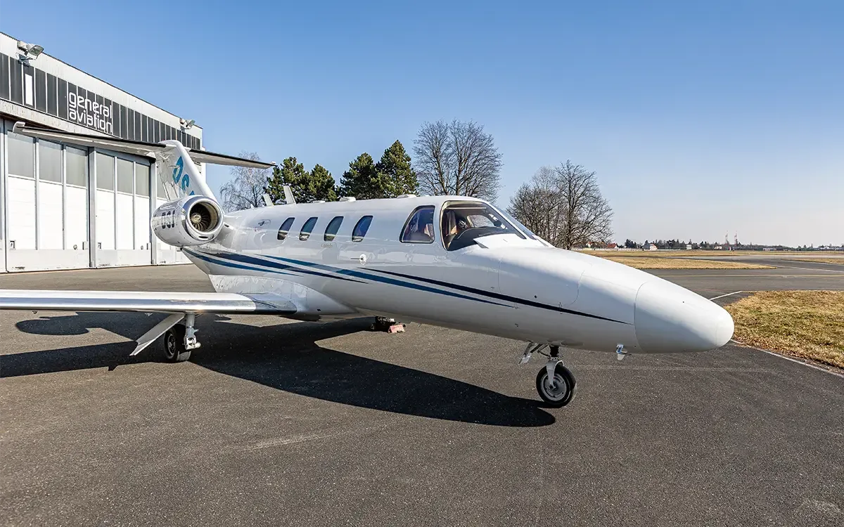 A white business jet with blue stripes is parked on a tarmac next to a 'general aviation' hangar under a clear blue sky.