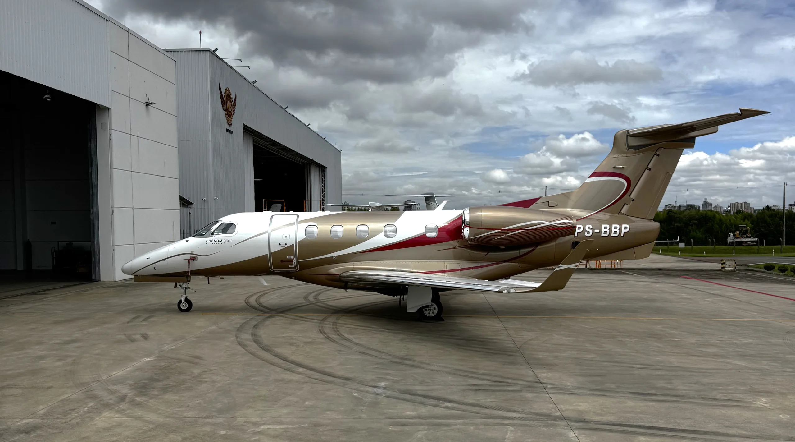 A gold and white Embraer Phenom 300 business jet with red accents is parked on an tarmac next to a hangar under a cloudy sky.