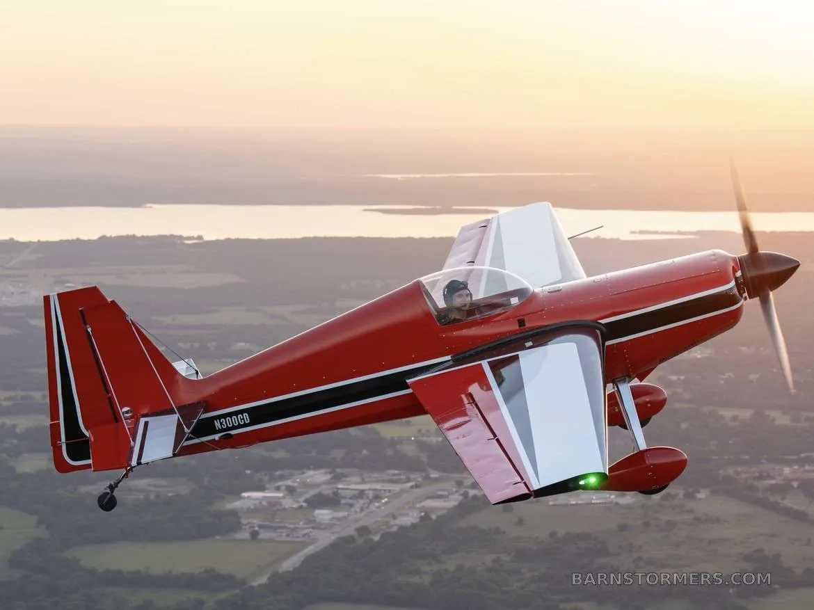 A red and white aerobatic aircraft, N300CB, with a pilot, flying over a landscape with a lake at sunset.