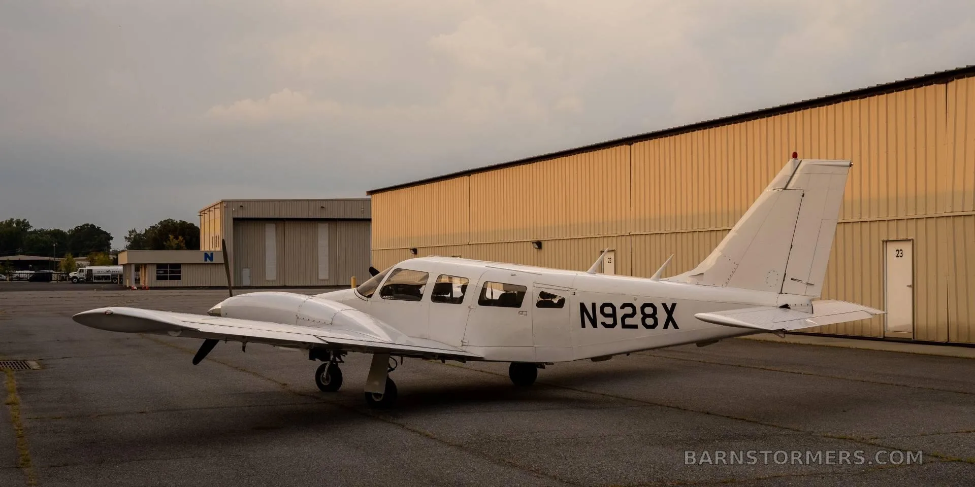 A white twin-engine propeller aircraft (N928X) is parked on an asphalt ramp with hangars in the background at sunset.