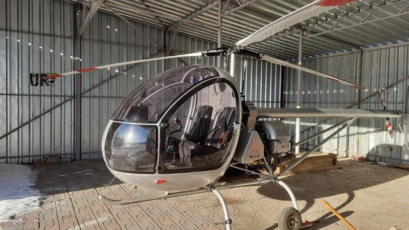 A silver AK1-3 light helicopter with a clear bubble canopy is parked inside a metal hangar.