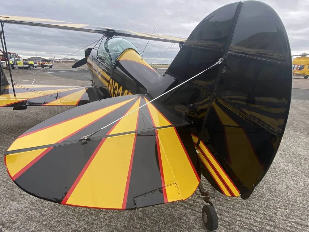 The black and yellow tail of the biplane N31475 is shown on an airfield with emergency vehicles in the background.