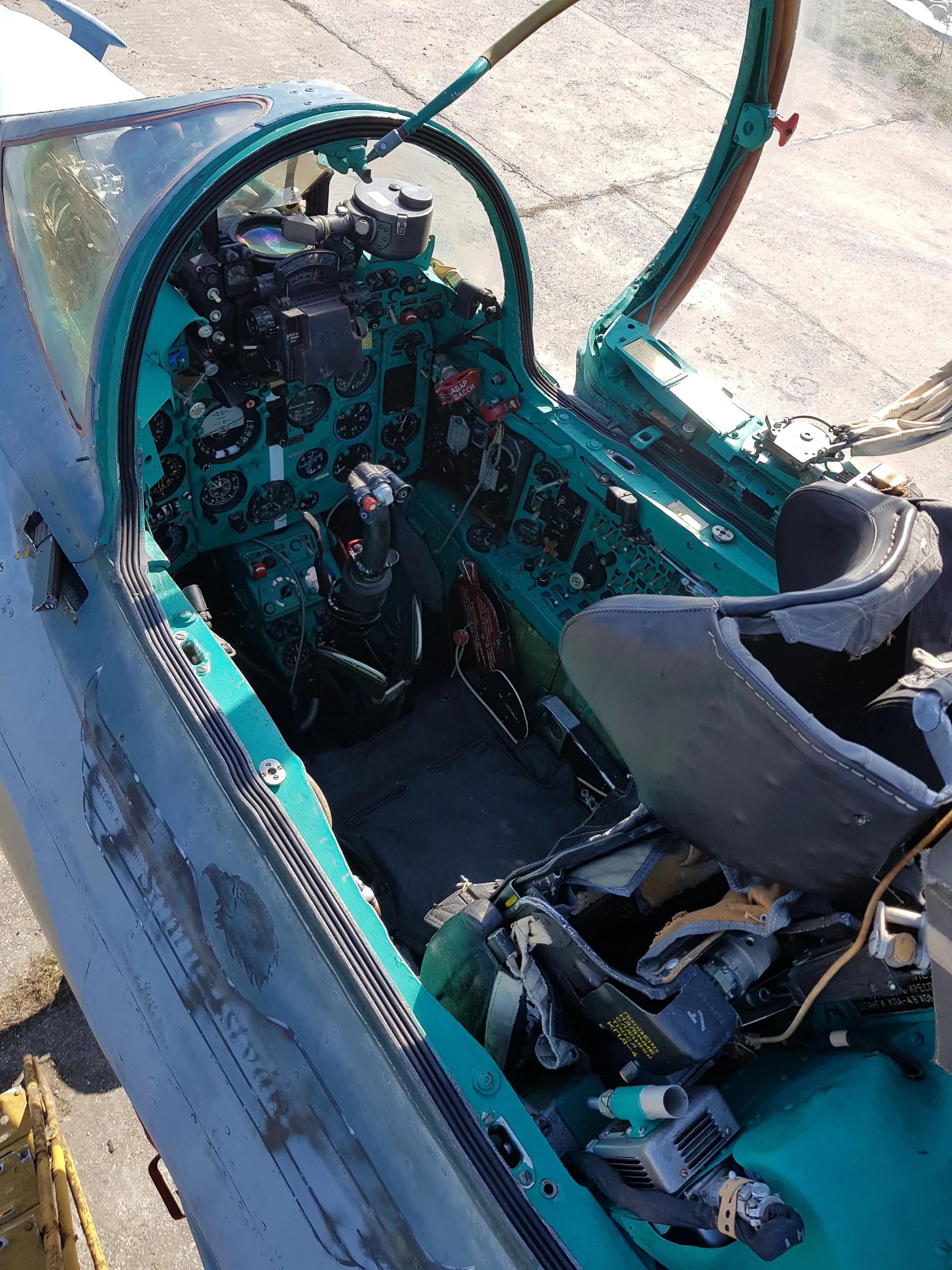An overhead view into the open cockpit of a MiG-21 fighter jet, revealing the green instrument panel, control stick, and ejection seat.
