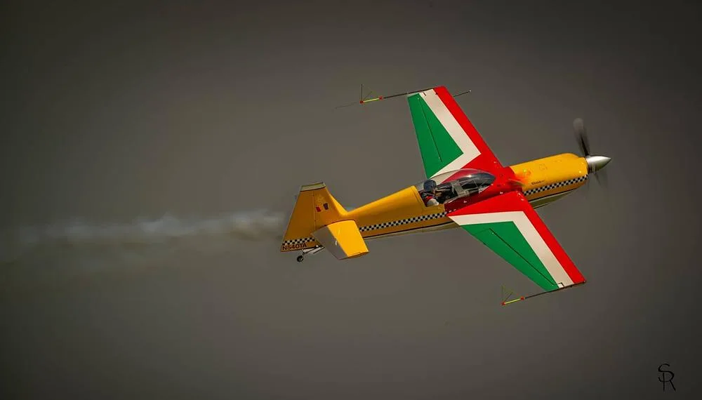 The N540TA aerobatic aircraft with red, green, yellow, and checkerboard livery flies in a cloudy sky.