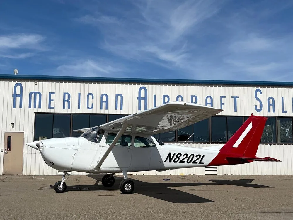 A white Cessna 172 with a red tail, registration N8202L, parked in front of a hangar under a blue sky.