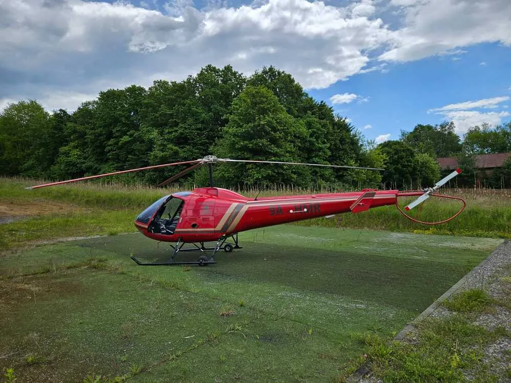 A red helicopter with gold stripes is parked on a green helipad, with a dense line of trees in the background under a partly cloudy sky.