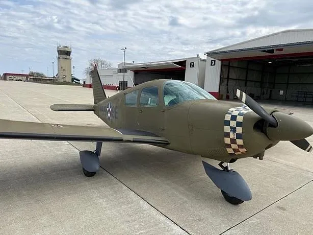 A green Grumman AA-5 Traveler aircraft with a checkered nose and US star insignia parked on an airfield with a control tower in the background.