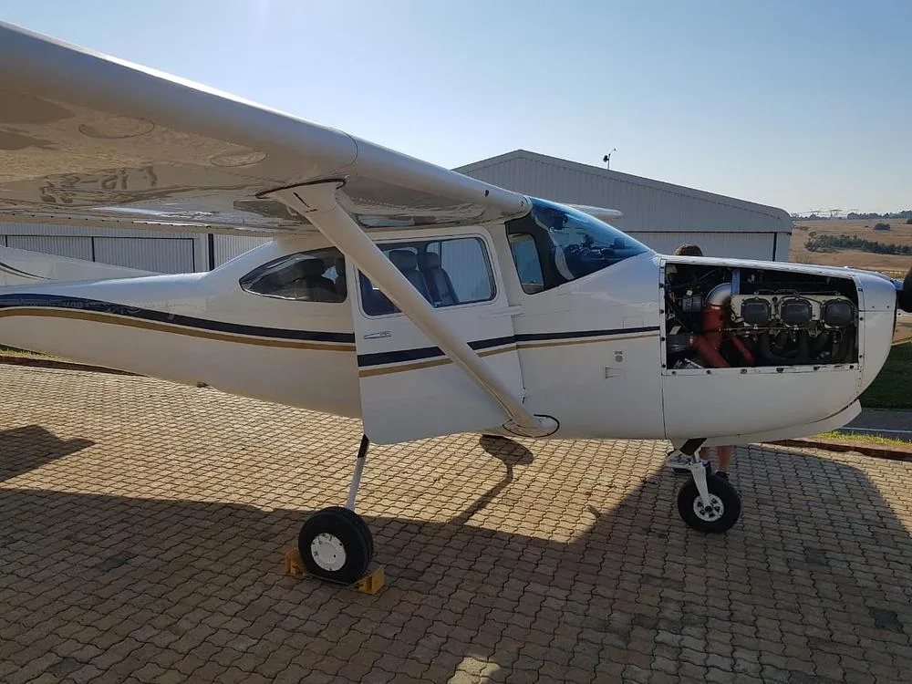 A white Cessna Skylane aircraft with blue and gold stripes parked outdoors with its engine cowling open, revealing the engine.