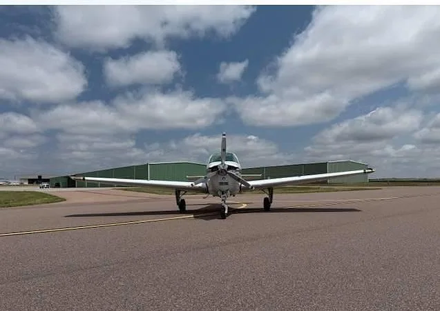 A white and gold Beechcraft Bonanza aircraft is seen from the front, parked on an airfield with hangars in the background.