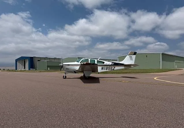 A white and gold Beechcraft Bonanza aircraft, tail number N1856Z, is parked on an airfield with green and blue hangars.