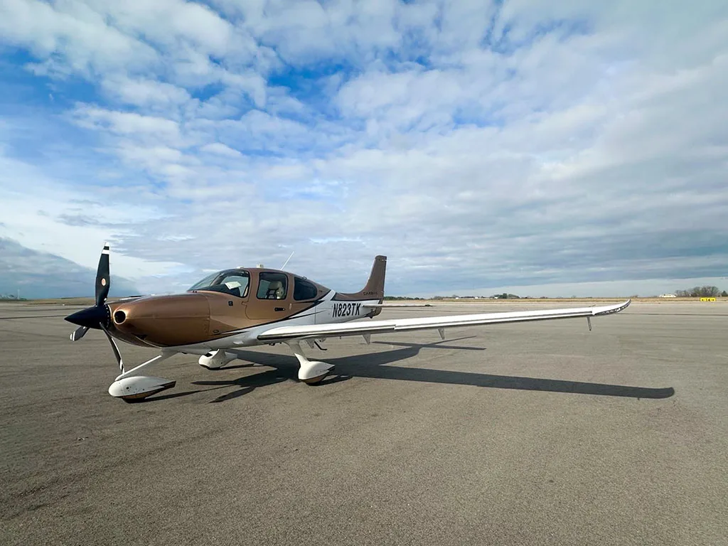 A Cirrus SR22 aircraft, N823TK, with a brown and white paint scheme parked on an airfield.