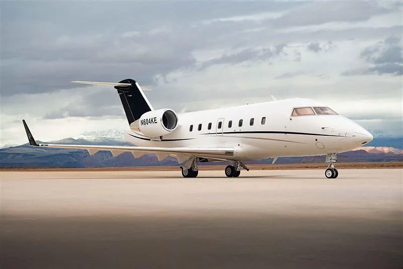 A white Bombardier Challenger 604 business jet with black accents, tail number N604KE, parked on an airfield under a cloudy sky.