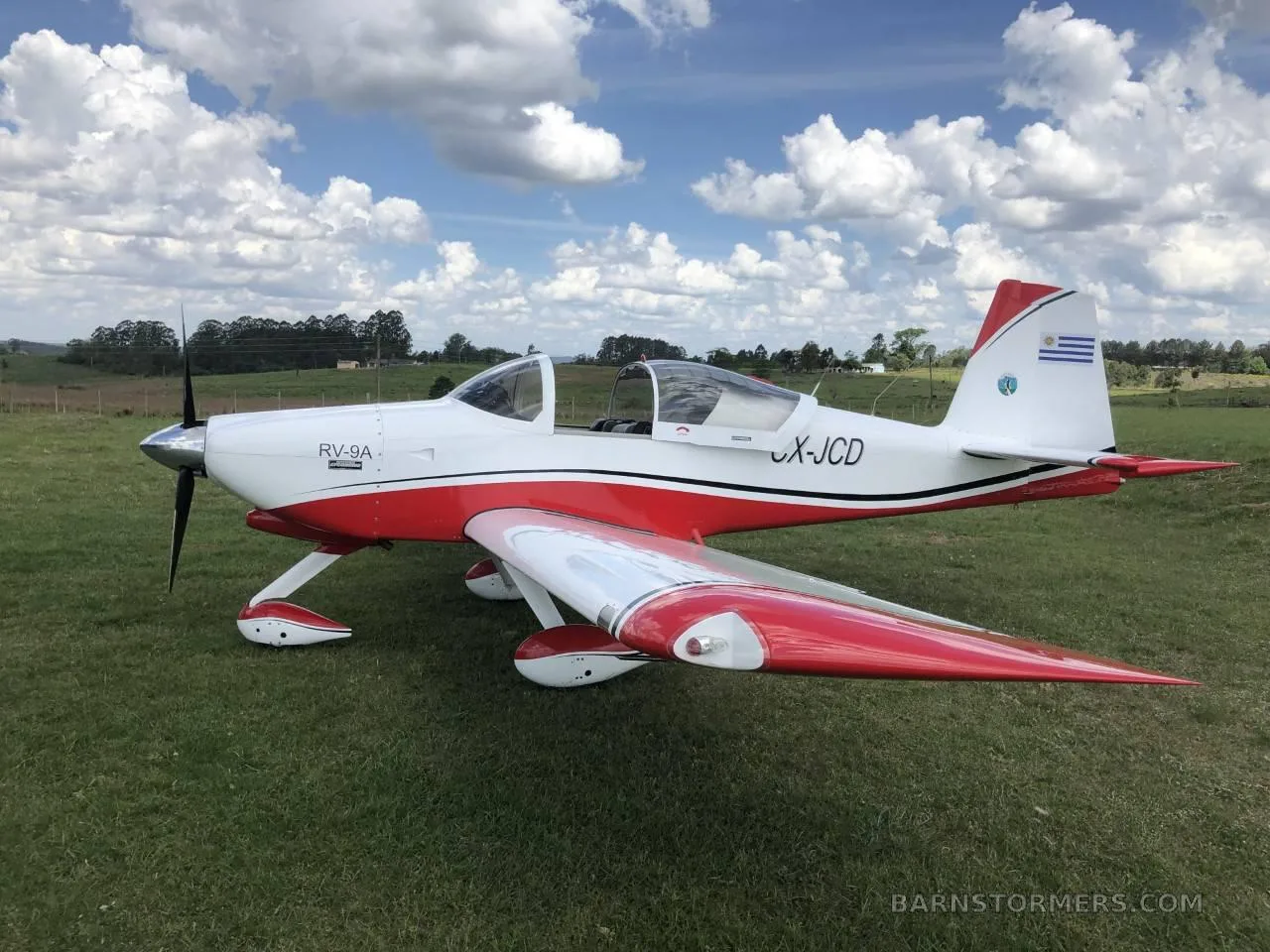 A white and red Van's RV-9A aircraft with Uruguayan flag on the tail, parked on a grassy field under a cloudy sky.