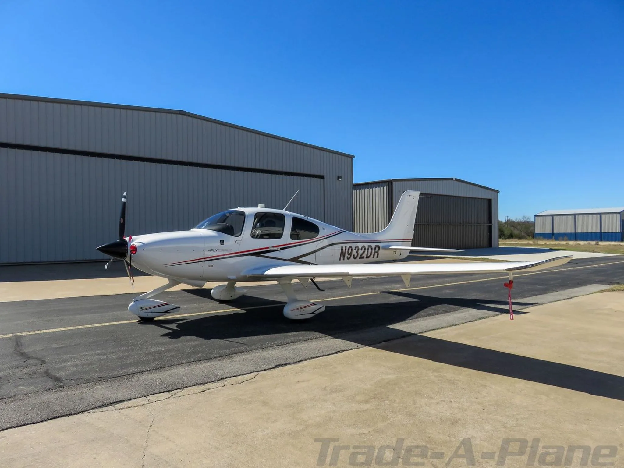 Side view of a white single-engine aircraft with red and black stripes, registration N932DR, parked on a tarmac in front of a hangar.