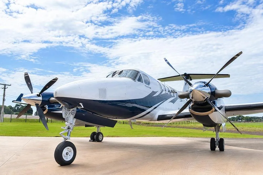 A front-quarter view of a white and blue Beechcraft King Air twin-turboprop aircraft with its propellers visible, parked on a tarmac.