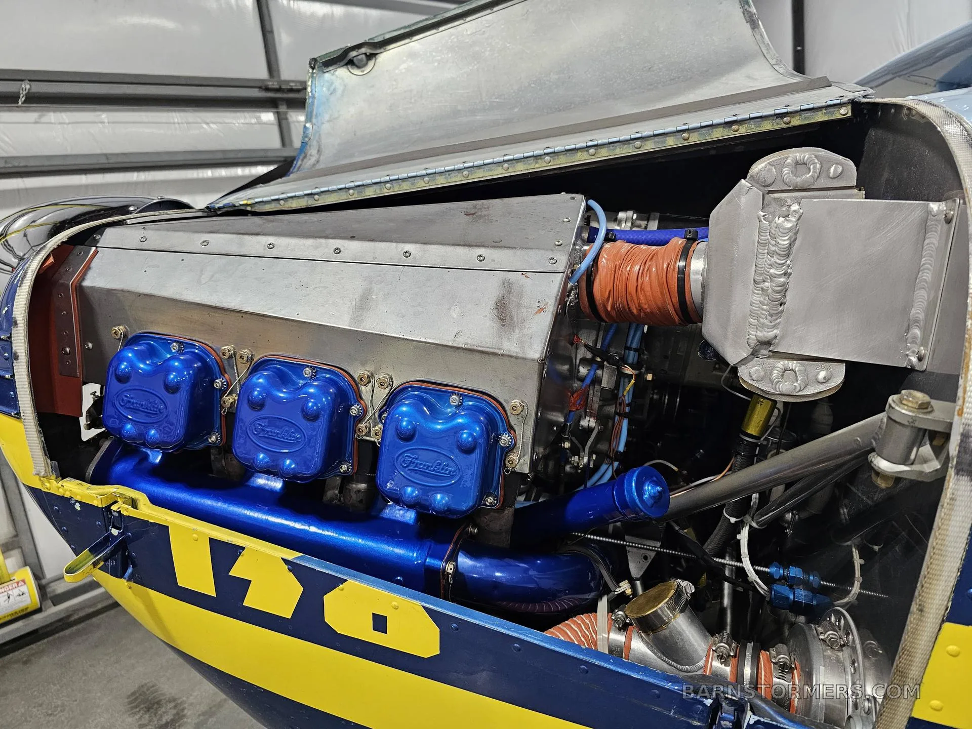 The open engine bay of an aircraft reveals a Franklin engine with blue valve covers and various mechanical components.