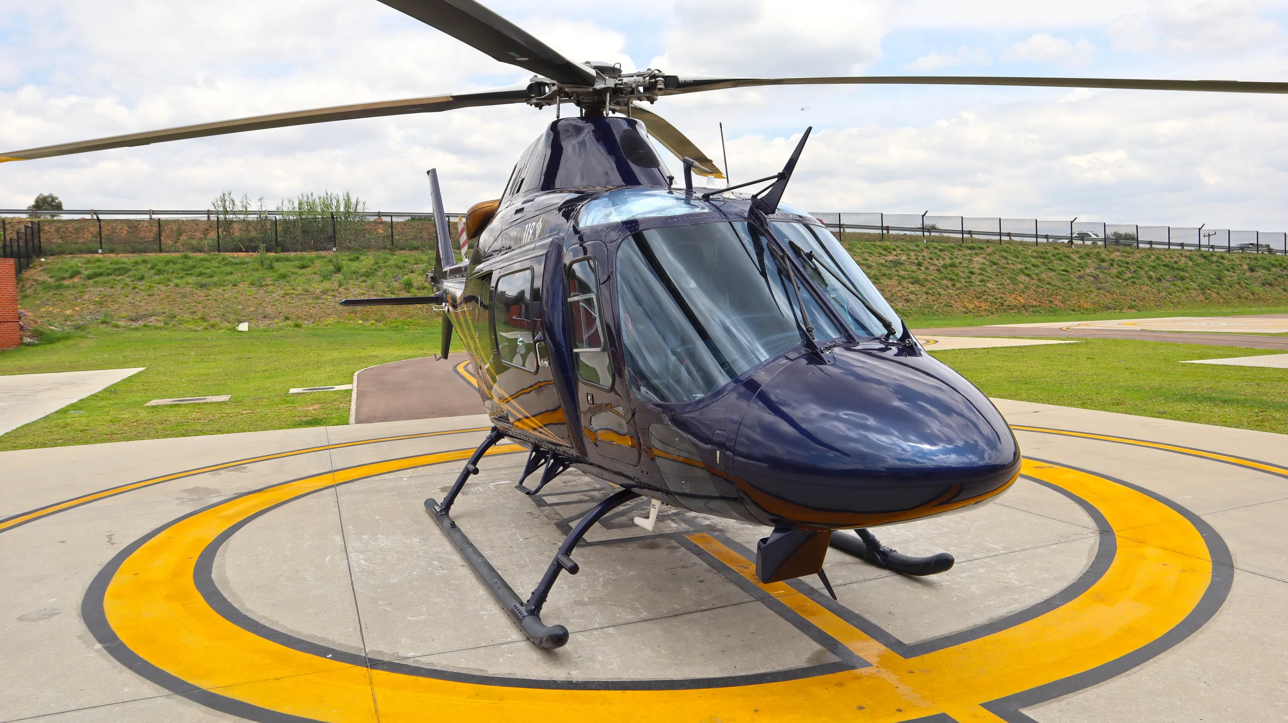A dark blue helicopter with a main rotor and tail rotor is parked on a concrete helipad with yellow markings.