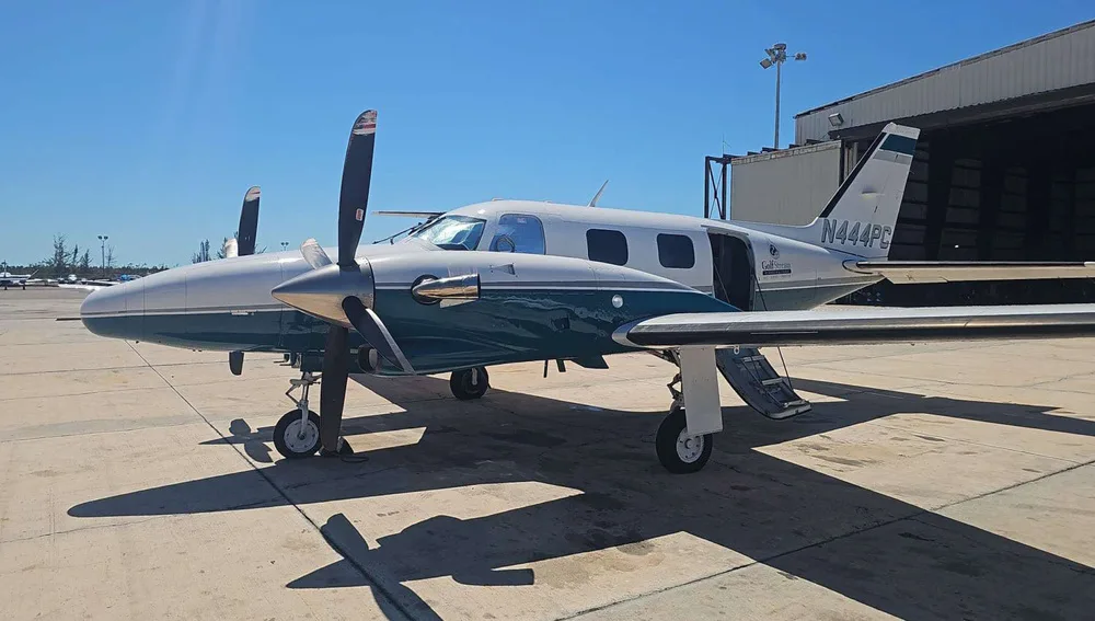 A Piper PA-31 Navajo, registration N444PC, in a white and teal paint scheme, parked on a tarmac with its passenger door open.