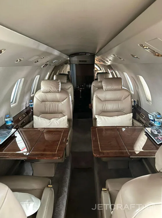 The interior cabin of a business jet, showing tan leather seats and wooden tables.