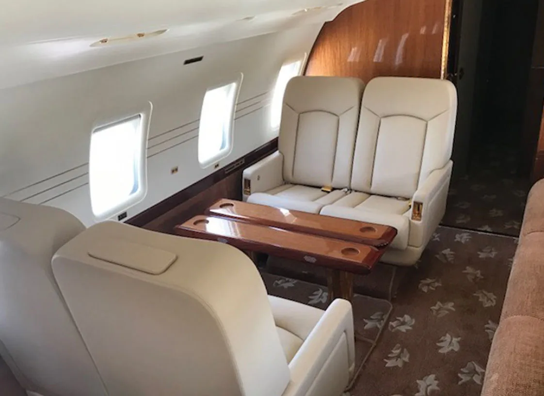 A private jet cabin features cream leather seats arranged around a wooden table with cup holders.