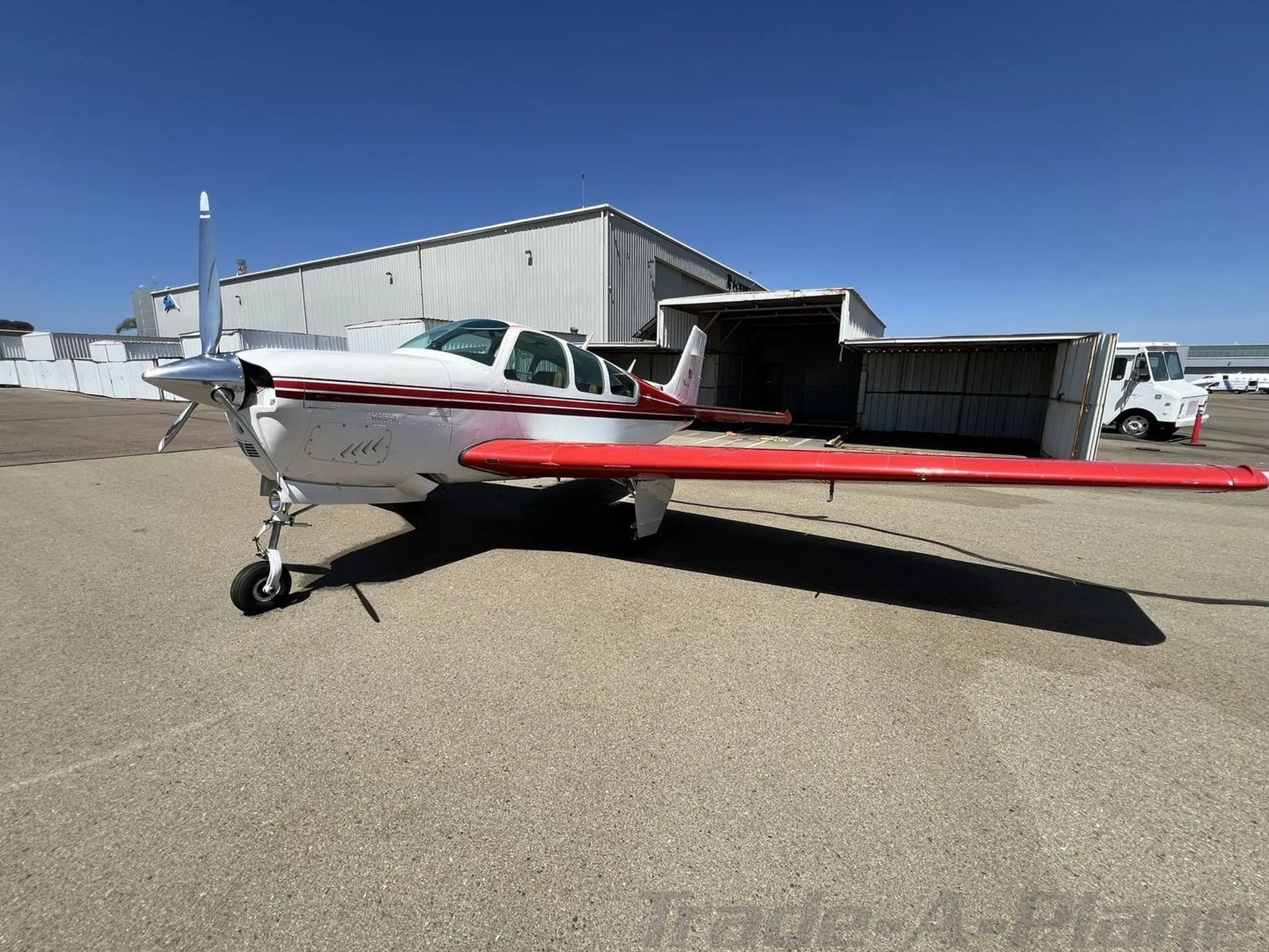 Three-quarter front view of a white and red Beechcraft Bonanza with a V-tail, parked on an airfield.