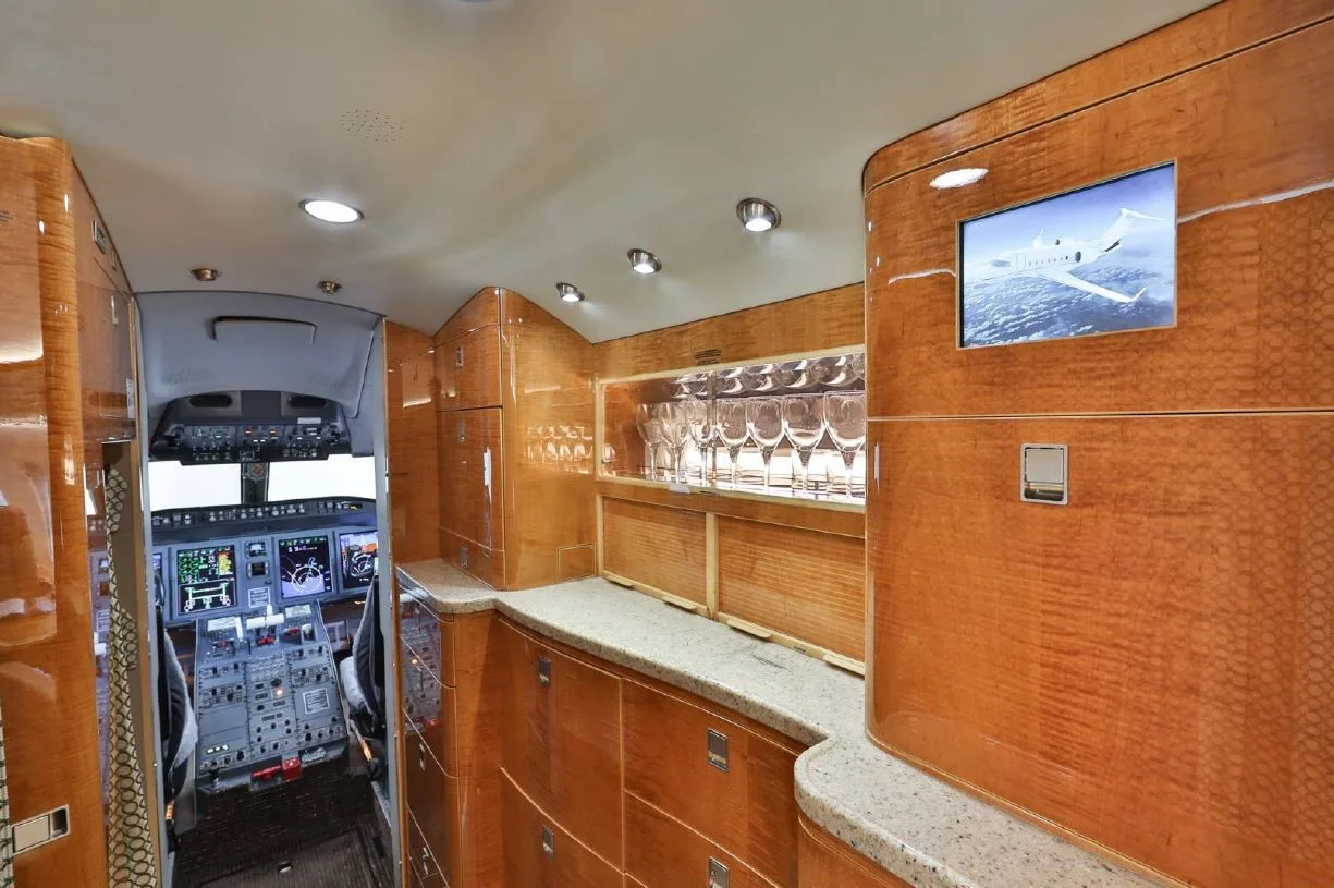 A luxurious galley with wood paneling and a screen showing an aircraft, with the cockpit visible in the background.