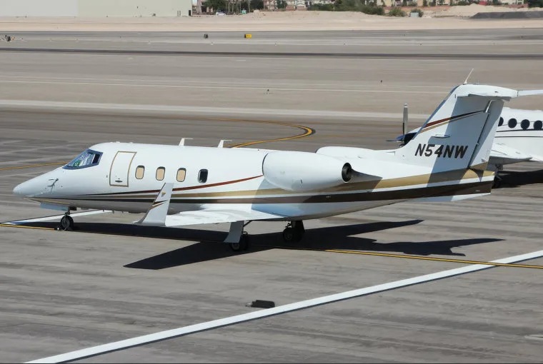 A white Learjet 60 with brown and gold stripes, registration N54NW, parked on an airport tarmac.