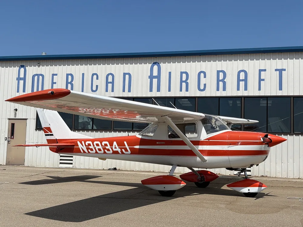 A side view of the red and white Cessna 172 aircraft N3834J parked on an airfield in front of a hangar.