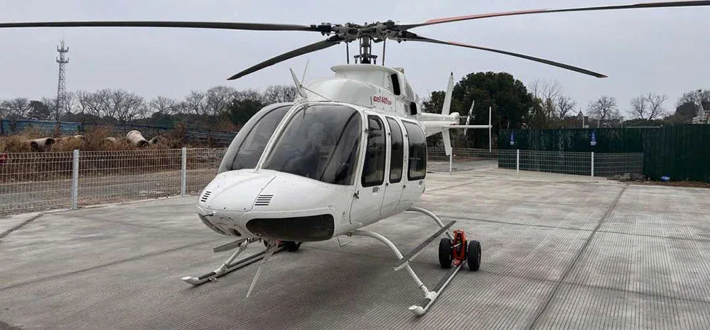 A white Bell 407GXP helicopter with landing skids and ground handling wheels, viewed from the front on a concrete pad.