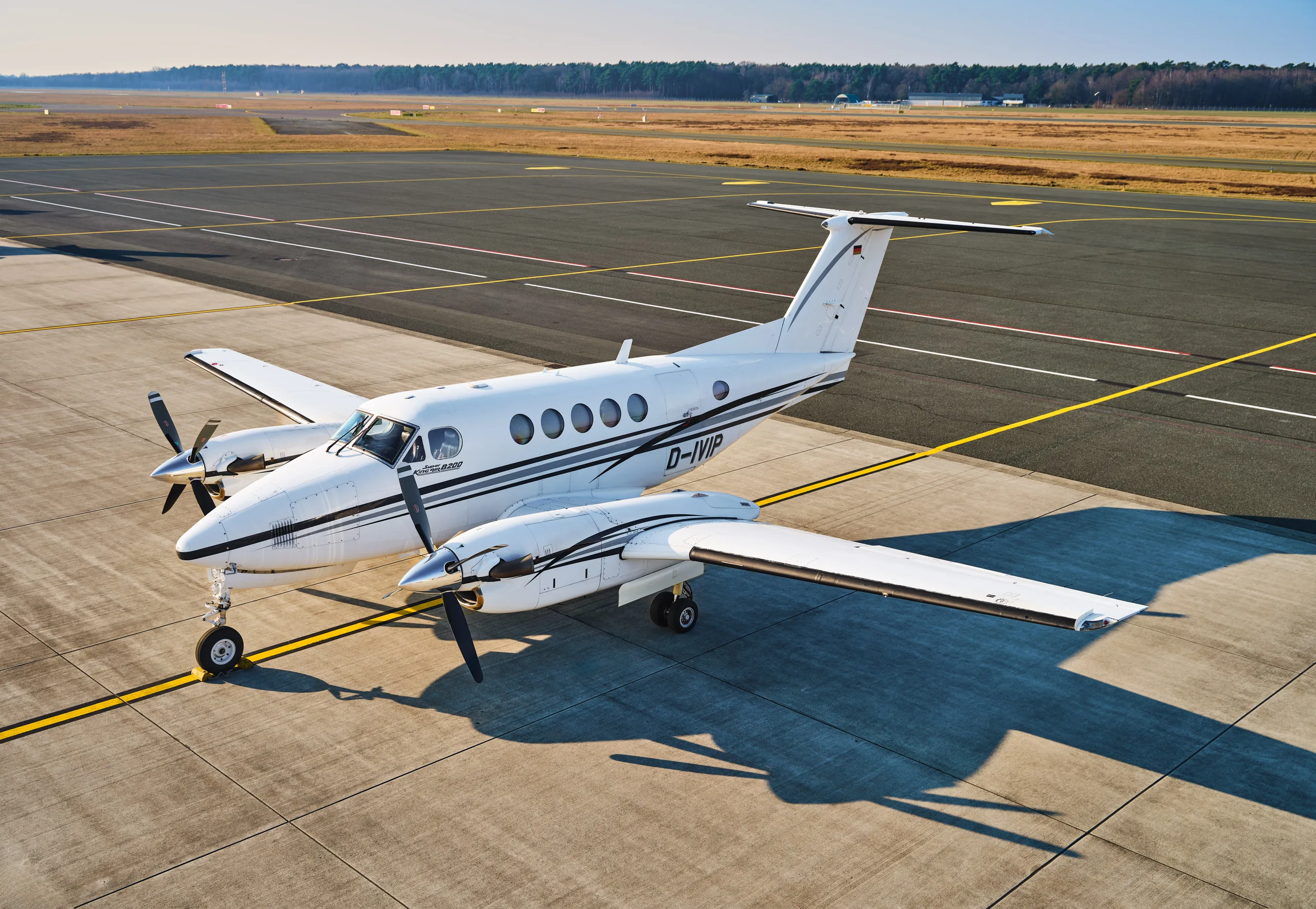 An aerial view of a white and black twin-turboprop aircraft, likely a King Air, parked on an airfield with long shadows.
