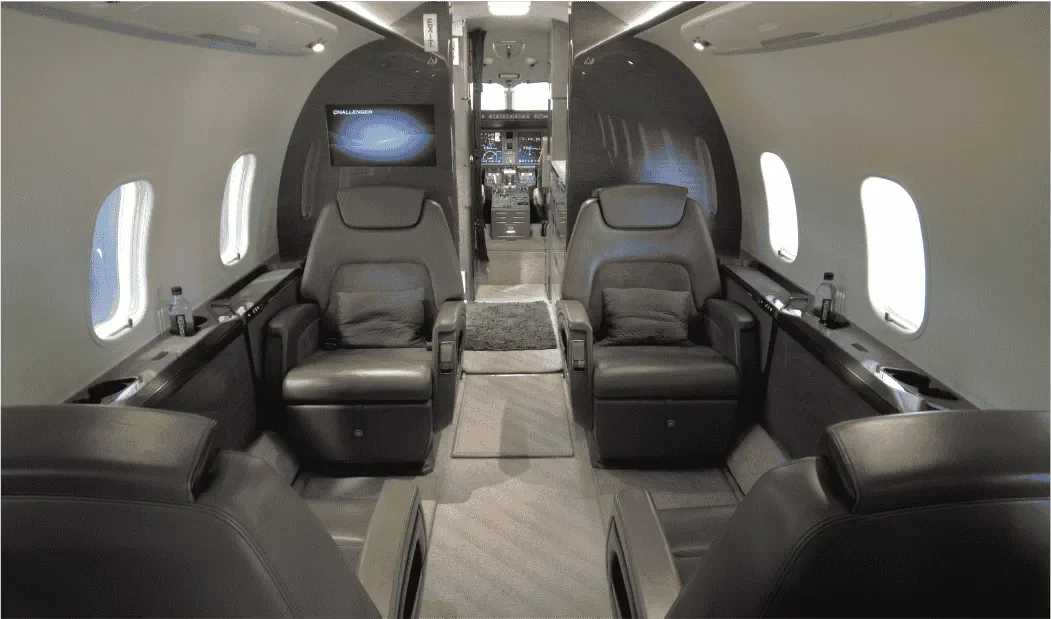 The luxurious interior of a business jet cabin with four grey leather seats facing each other, and the cockpit visible in the background.