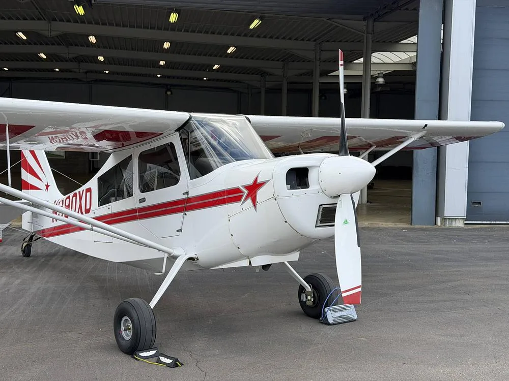 A white single-engine aircraft with red stripes and a red star on the nose, parked on an asphalt apron in front of a hangar.