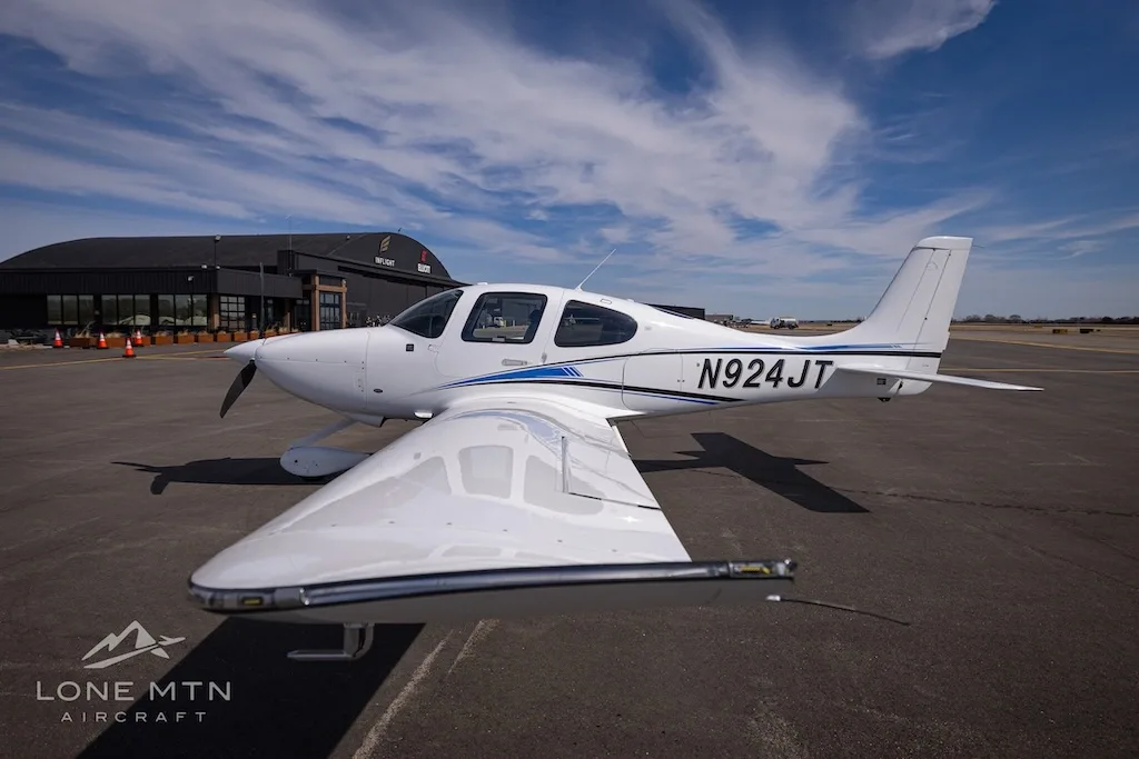 A white single-engine aircraft with blue and black stripes, tail number N924JT, parked on an airport tarmac with a building in the background.