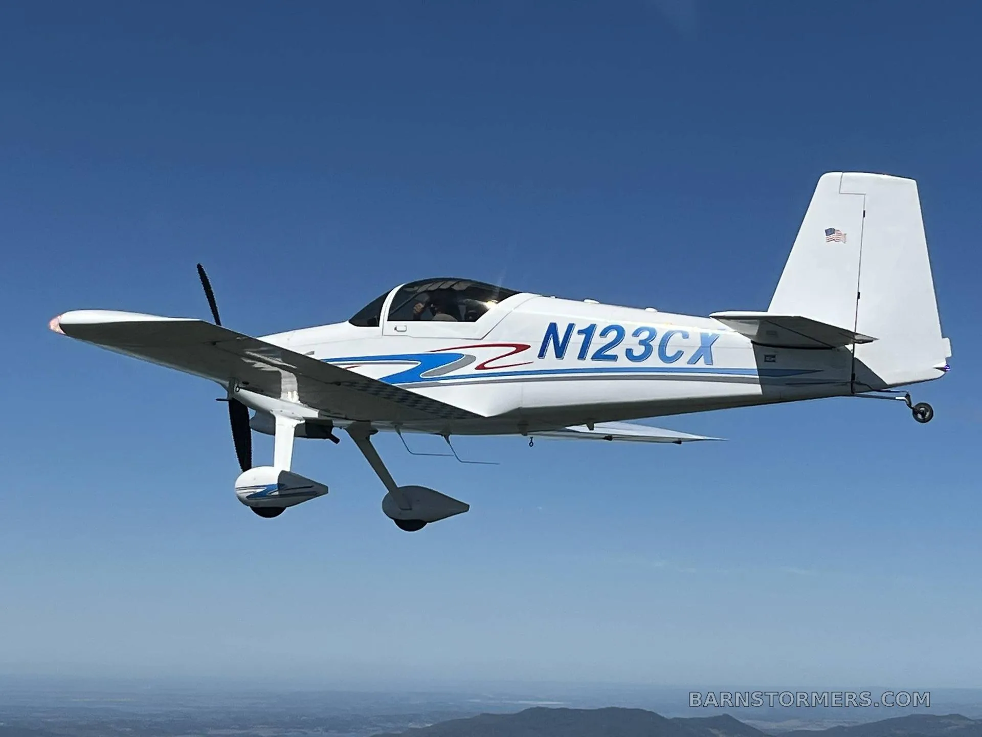 A white single-engine aircraft with blue and red stripes (N123CX) flying against a clear blue sky over mountains.