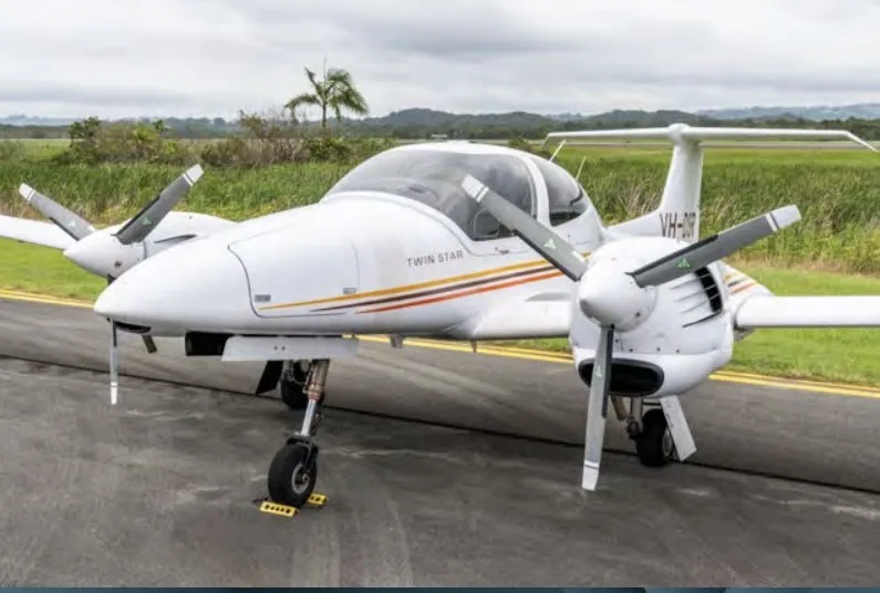 A white Diamond DA42 Twin Star aircraft with registration VH-DIR is parked on an asphalt runway with green fields in the background.