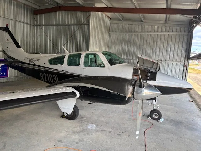 A white and dark grey twin-engine aircraft, N2103L, with its engine cowling open, parked in a hangar.