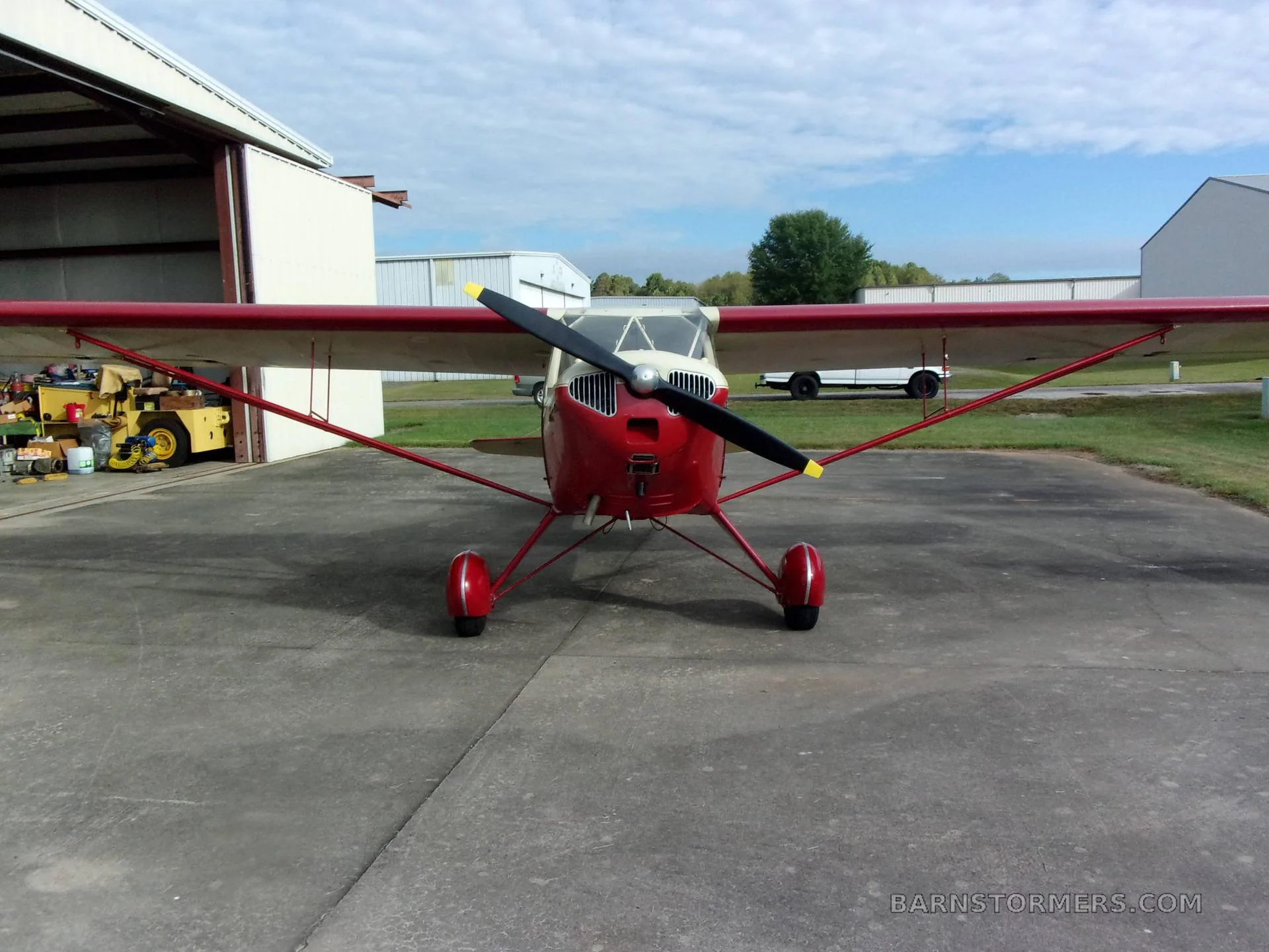 A red and cream Taylorcraft BC-12D aircraft parked on a concrete apron outside a hangar, viewed from the front.