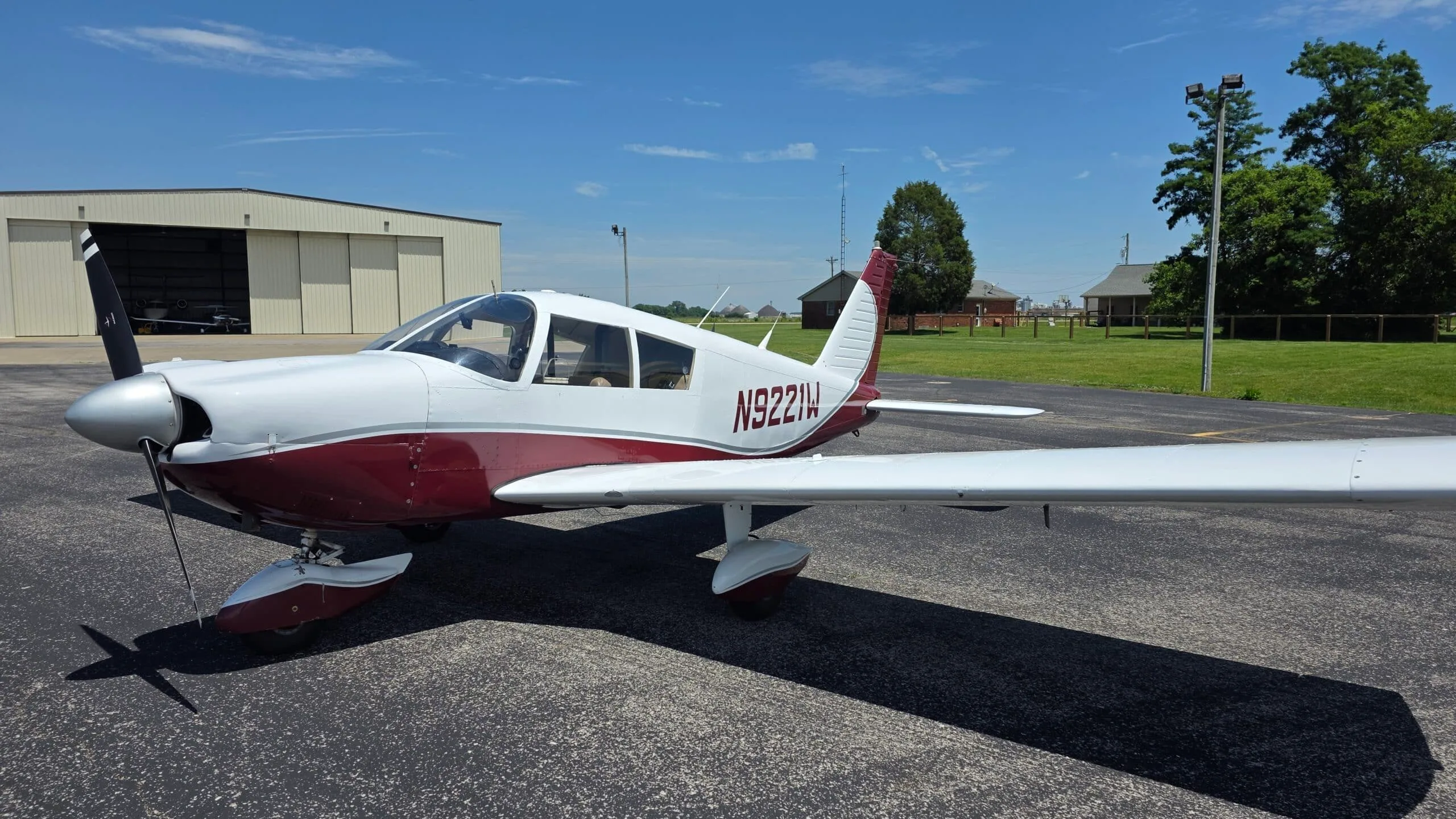 A white and maroon Piper Cherokee (N9221W) parked on an airfield with hangars in the background.