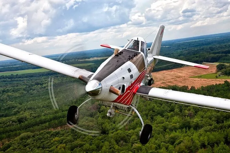 A white, red, and black agricultural aircraft flies over a green forest under a cloudy sky.