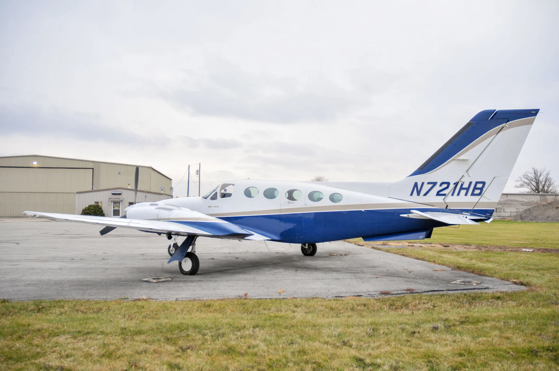 A side-rear view of the N721HB aircraft, a white and blue twin-engine plane, parked on an airfield.