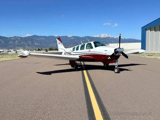 A white and red Beechcraft Bonanza aircraft is parked on an asphalt runway with mountains in the background.